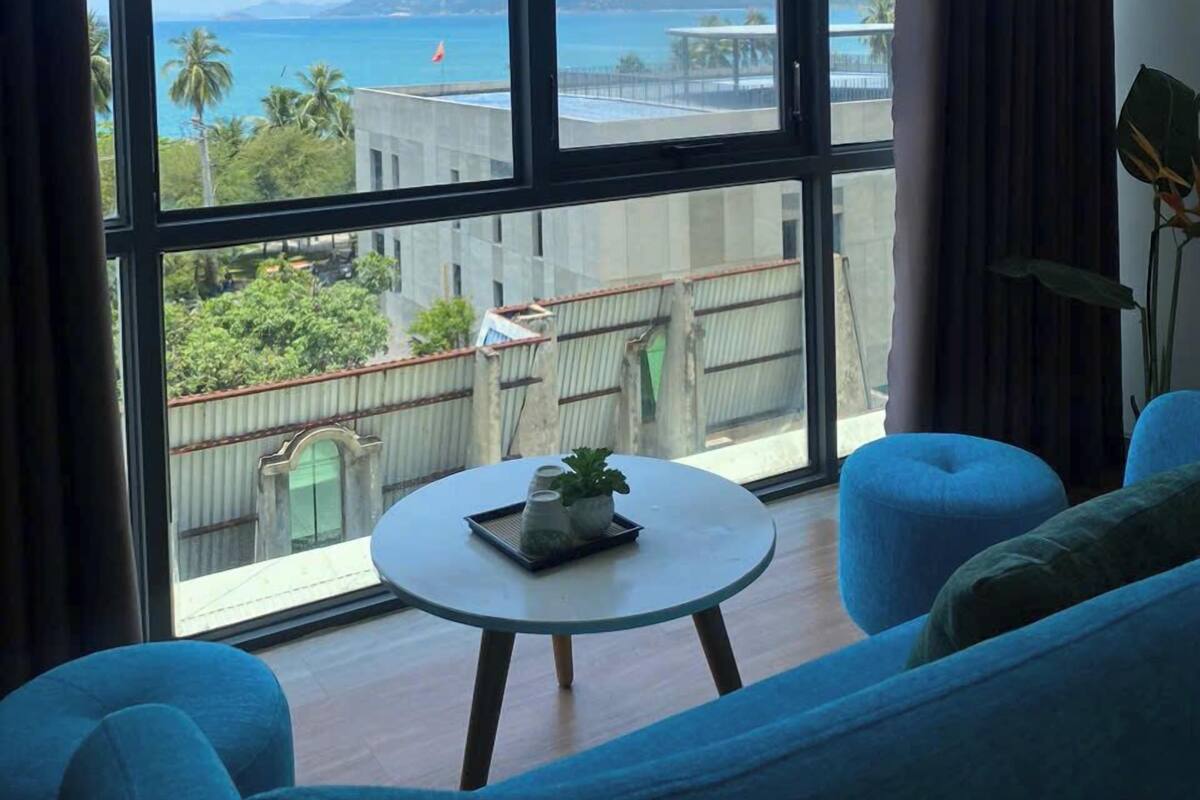 Habitación con amplia ventana que ofrece vistas a la playa y las montañas. Se observa una mesa redonda con una planta y un libro, además de mobiliario tapizado en azul.