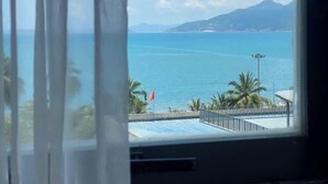 A view through a window with sheer curtains, featuring a coastal landscape with a clear sky, a flag, and palm trees. - The H House Quy Nhơn (Quy Nhon)