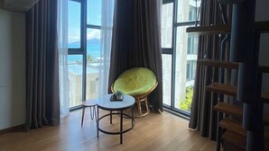 A spacious room with a large window offering a view of the sea, a round table with a chair, and a staircase with a railing. - The H House Quy Nhơn (Quy Nhon)