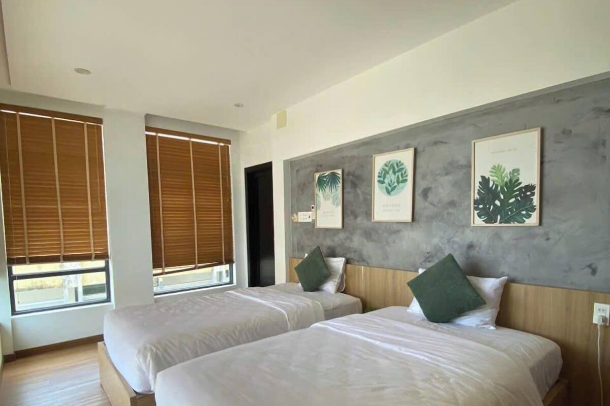 Una habitación de hotel moderna con dos camas, suelo de madera, ventanas amplias con persianas e impresiones botánicas enmarcadas en la pared.