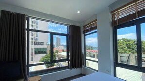 A hotel room with a large bed, two windows with curtains, a small table, and a view of the cityscape. - The H House Quy Nhơn (Quy Nhon)