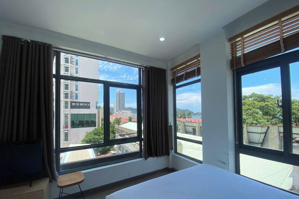 Habitación de hotel con una cama grande, dos ventanas con cortinas, una mesita y vistas a la ciudad.