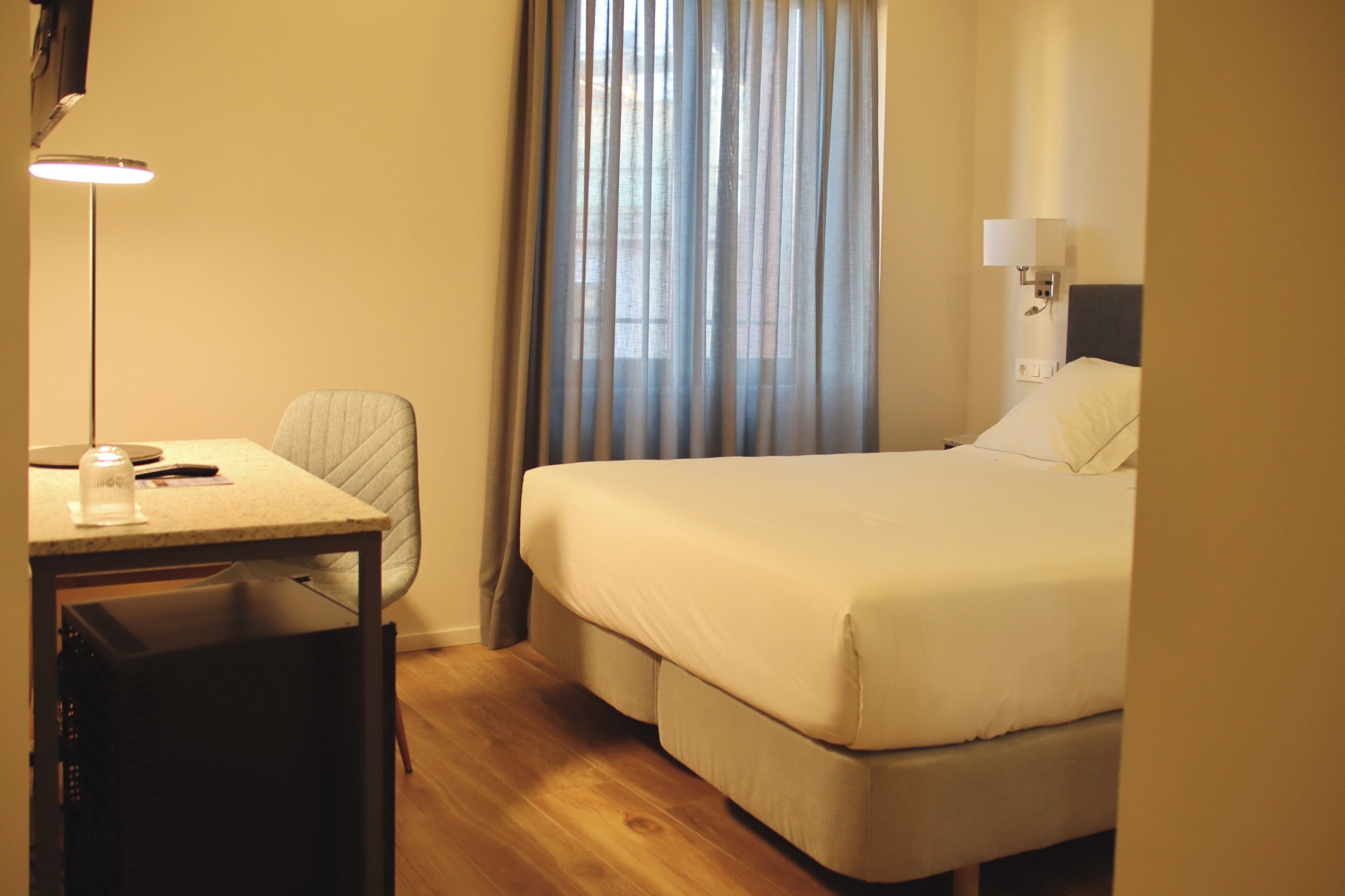 Una habitación de hotel con cama, escritorio, silla, lámpara y ventana con cortinas.