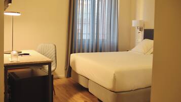 Una habitación de hotel con cama, escritorio, silla, lámpara y ventana con cortinas.