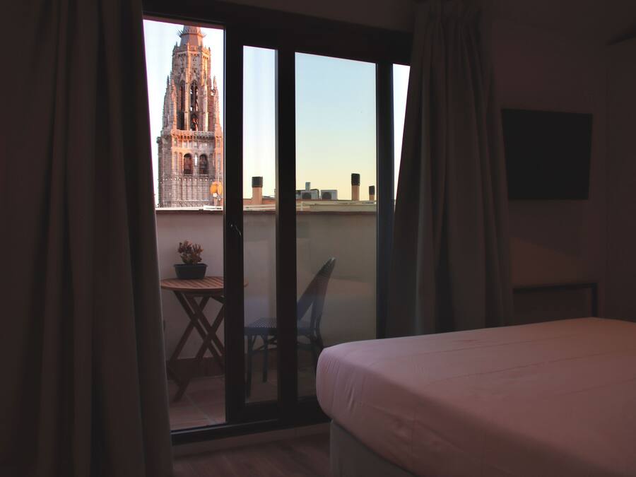 A room with a bed, a chair, a small table with a plant, and a view of a building through a sliding door.