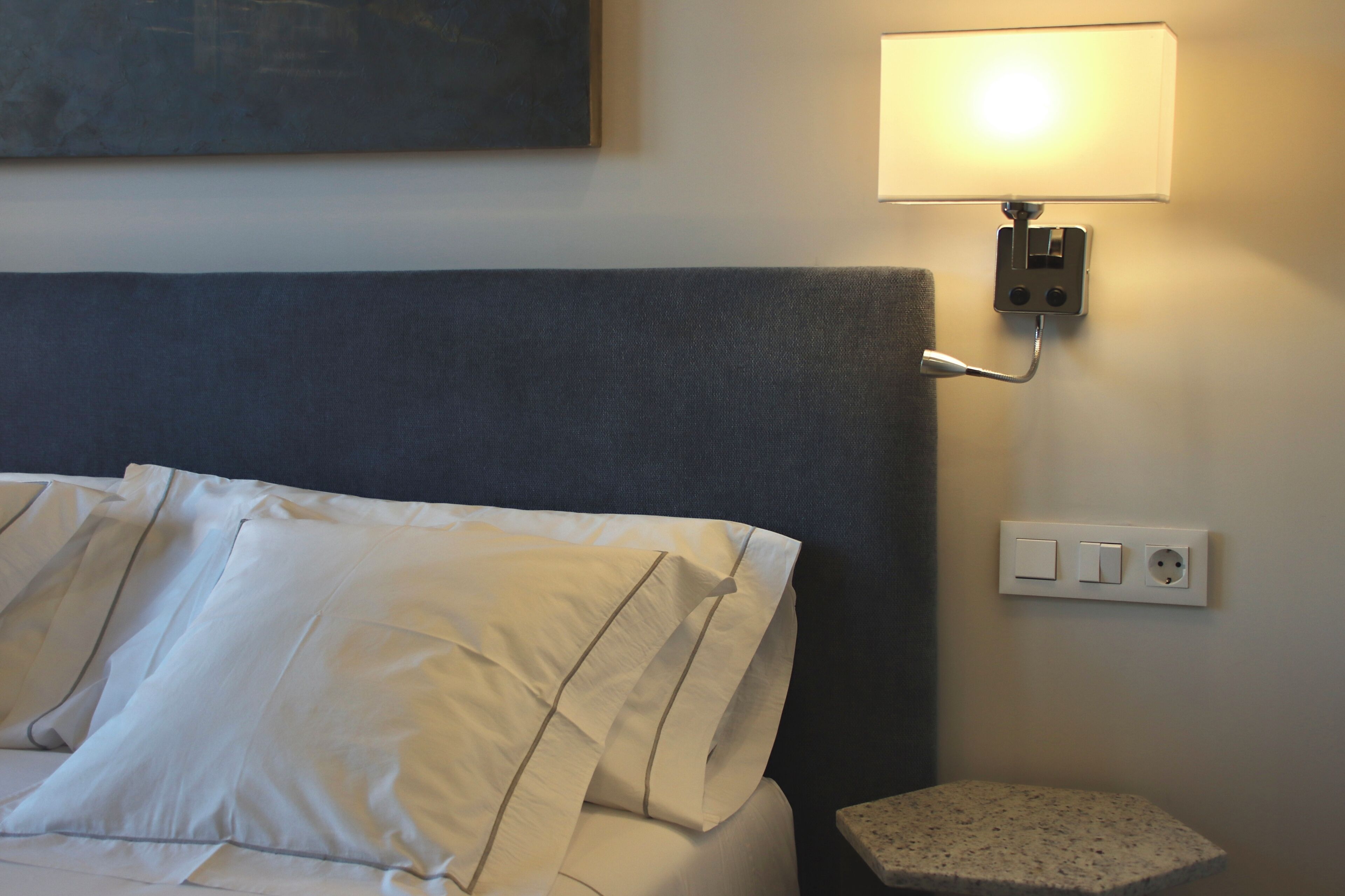 Una cama con ropa de cama blanca, cabecera gris, lámpara de pared y toma de corriente.