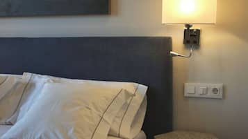 Una cama con ropa de cama blanca, cabecera gris, lámpara de pared y toma de corriente.