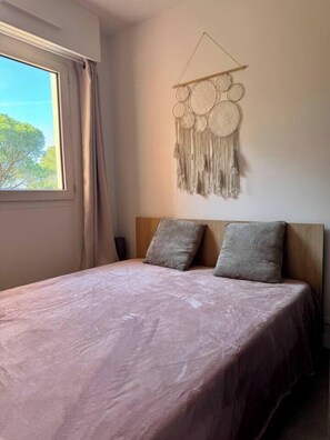 Un dormitorio con una cama, dos almohadas, un adorno de macramé en la pared y una ventana con cortinas.