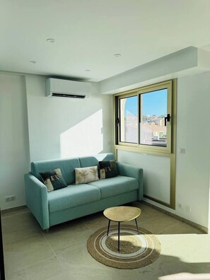 Uma sala de estar moderna com um sofá azul claro, uma mesa redonda pequena e uma janela grande com vista para a cidade.