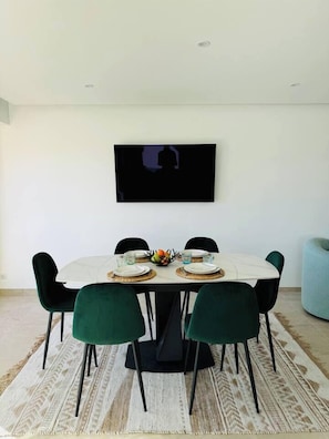 Uma sala de jantar com uma mesa branca, cadeiras verdes e uma televisão de tela plana na parede.