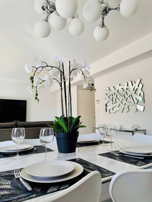 Uma sala de jantar moderna com uma mesa branca posta para quatro pessoas, uma orquídea em vaso e uma obra de arte geométrica na parede.