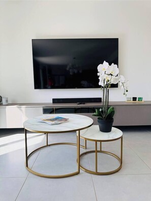 Uma sala de estar moderna com uma grande televisão de tela plana na parede, duas mesas redondas com molduras douradas, uma orquídea em vaso e uma revista sobre uma das mesas.