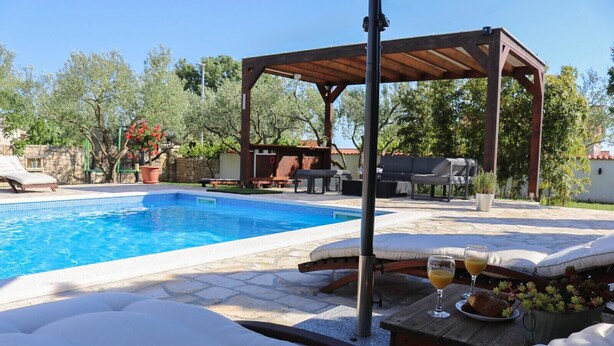 Ein Poolbereich mit einer Pergola, Liegestühlen und einem Tisch mit einem Glas Orangensaft.