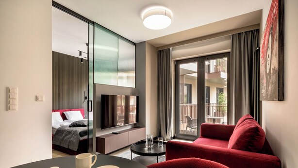 Un hotel moderno con cama, sofá rojo, televisión, mesa de comedor con un plato blanco y una taza, y balcón con vistas.