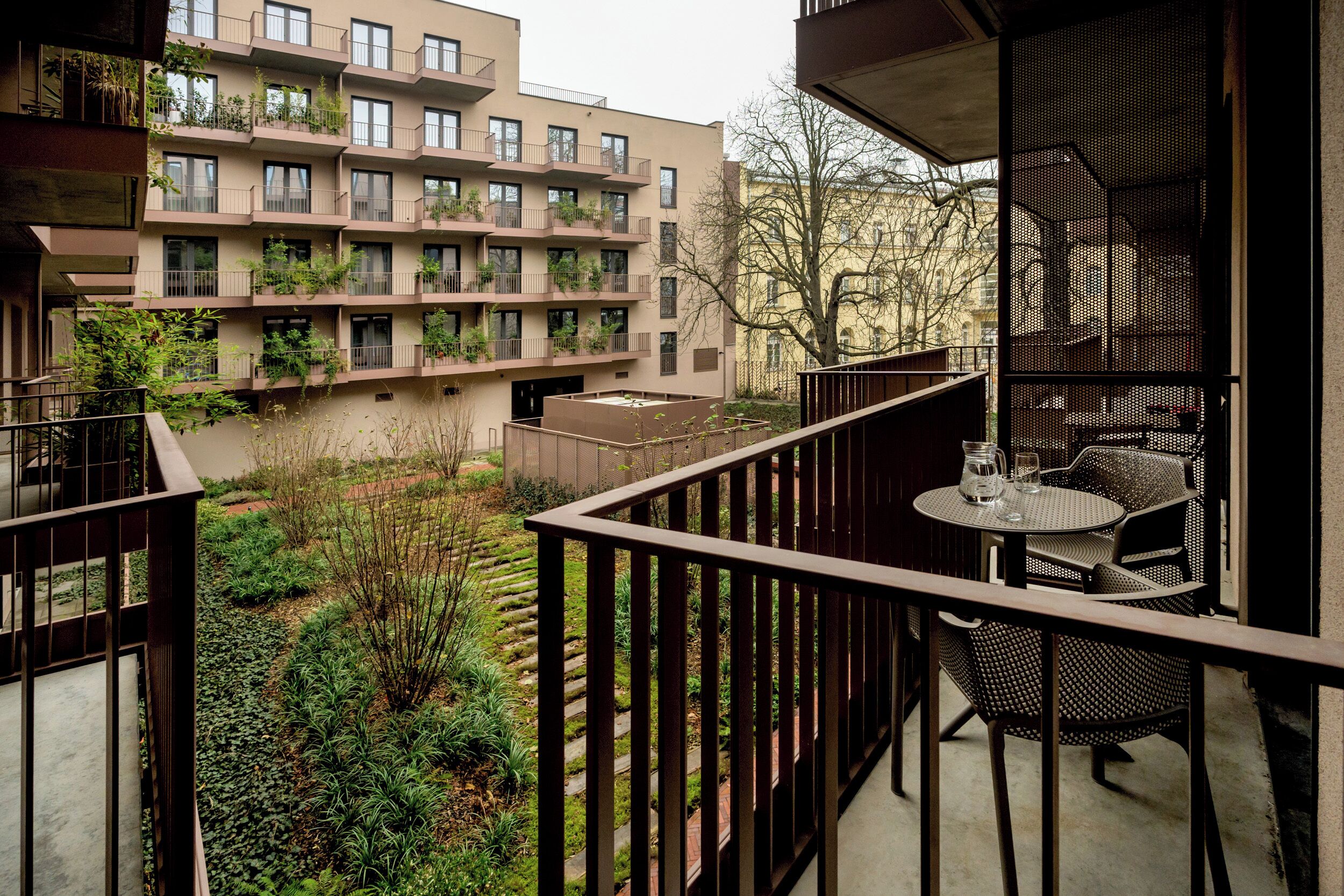 Ein Balkon mit Tisch und Stühlen, umgeben von viel Grün und Blick auf ein mehrstöckiges Gebäude mit Balkonen.