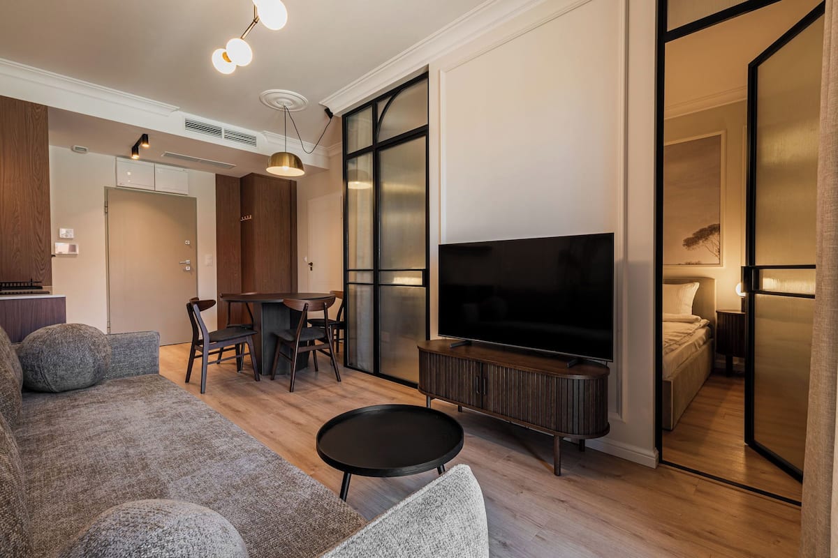 A modern living room with a sofa, a TV, a dining area, and a bedroom visible through a sliding door.