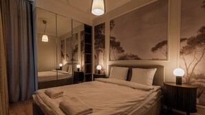 Una cama bien hecha con cabecera, lámparas de noche, un espejo grande y un cuadro de árboles en la pared.