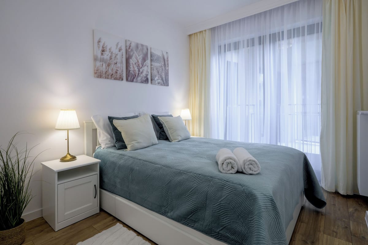 A neatly made bed with a blue bedspread, two bedside lamps, and a plant on the left.