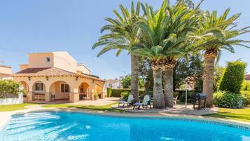 A swimming pool with lounge chairs, palm trees, and a Mediterranean-style building with arches.