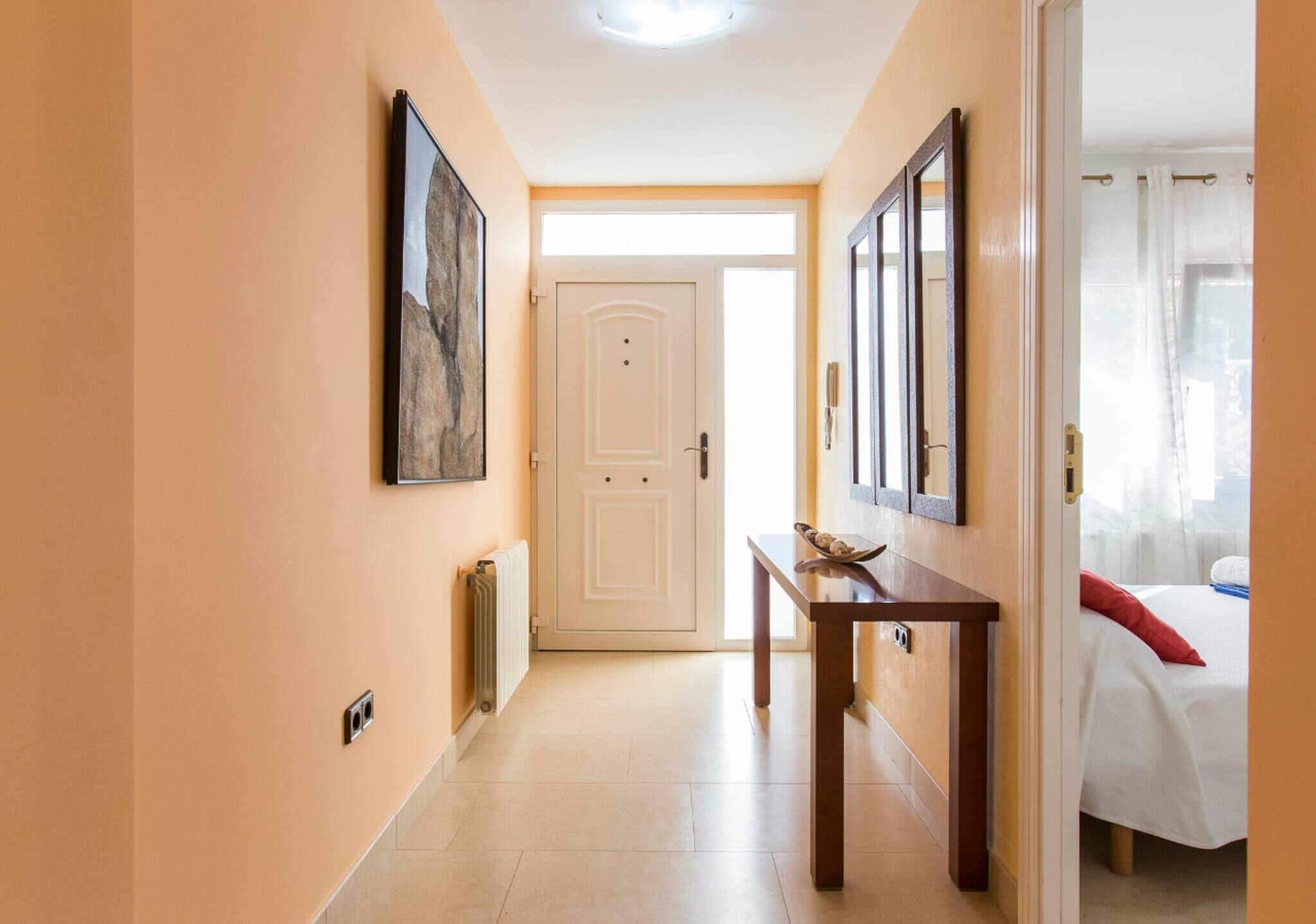 A hallway with a white door, a table with a bowl, and a bed with a red pillow.
