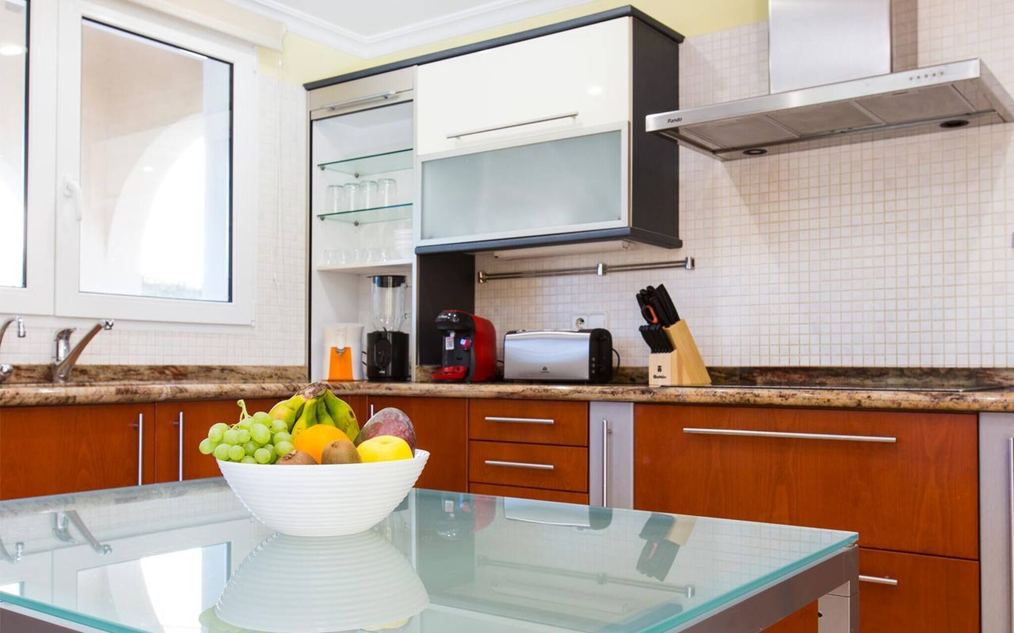 A modern kitchen with wooden cabinets, a glass-top table, a bowl of fruit, a coffee maker, and a toaster.