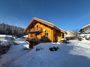 A wooden chalet with a balcony, surrounded by snow-covered ground and trees.