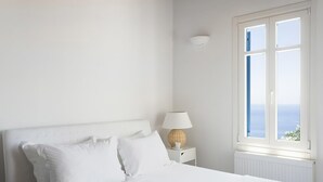 A white bedroom with a bed, a nightstand with a lamp, and a window with a view of the sea. - My Rental Homes - Villa Elpida with infinity pool overlooking  the Aegean sea (Mykonos)