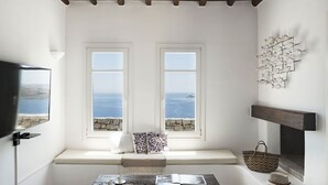A room with a stone bench by two windows overlooking the sea, a fireplace, and a wall-mounted fish decoration. - My Rental Homes - Villa Elpida with infinity pool overlooking  the Aegean sea (Mykonos)