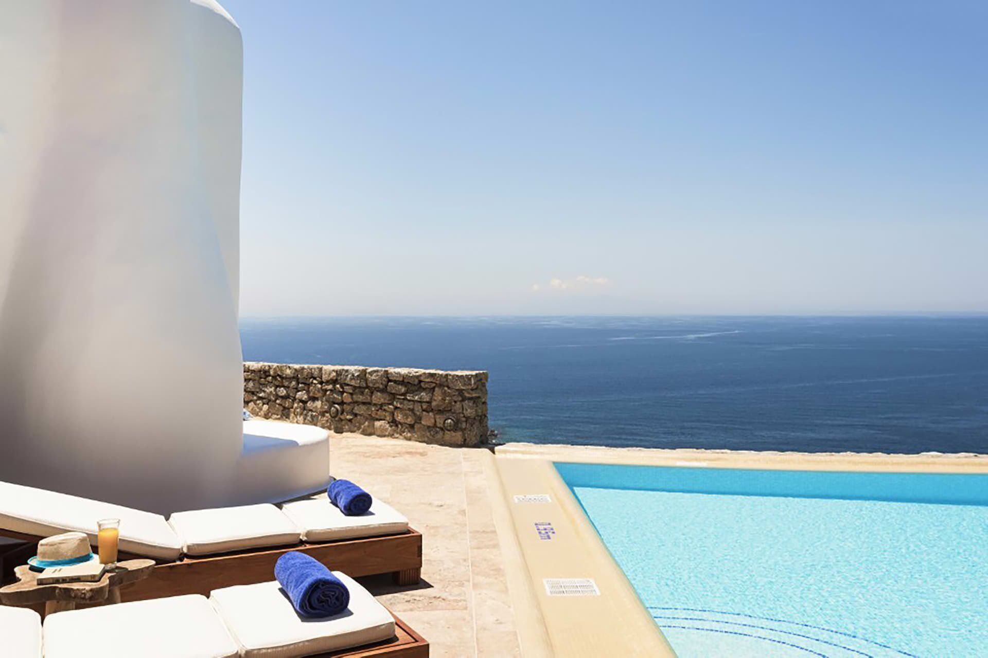 Un espace aménagé au bord de la piscine, avec des chaises longues et un mur en pierre, offrant une vue sur une mer d’un bleu limpide.