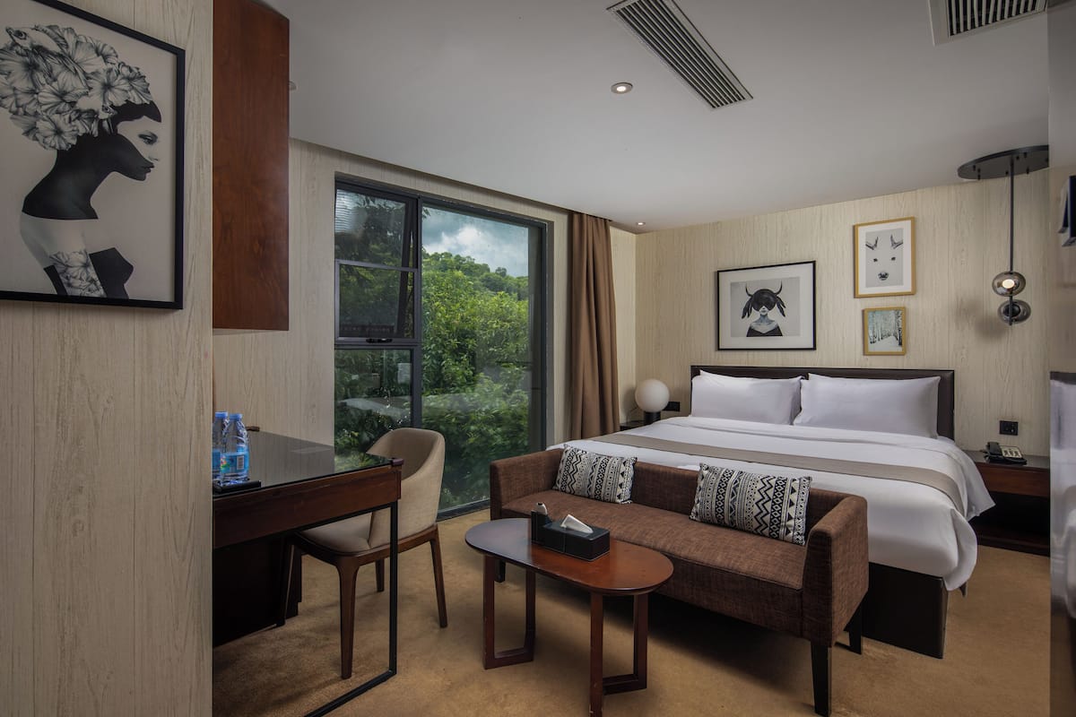 A hotel room with a large bed, a sofa, a desk, and a view of greenery through the window.