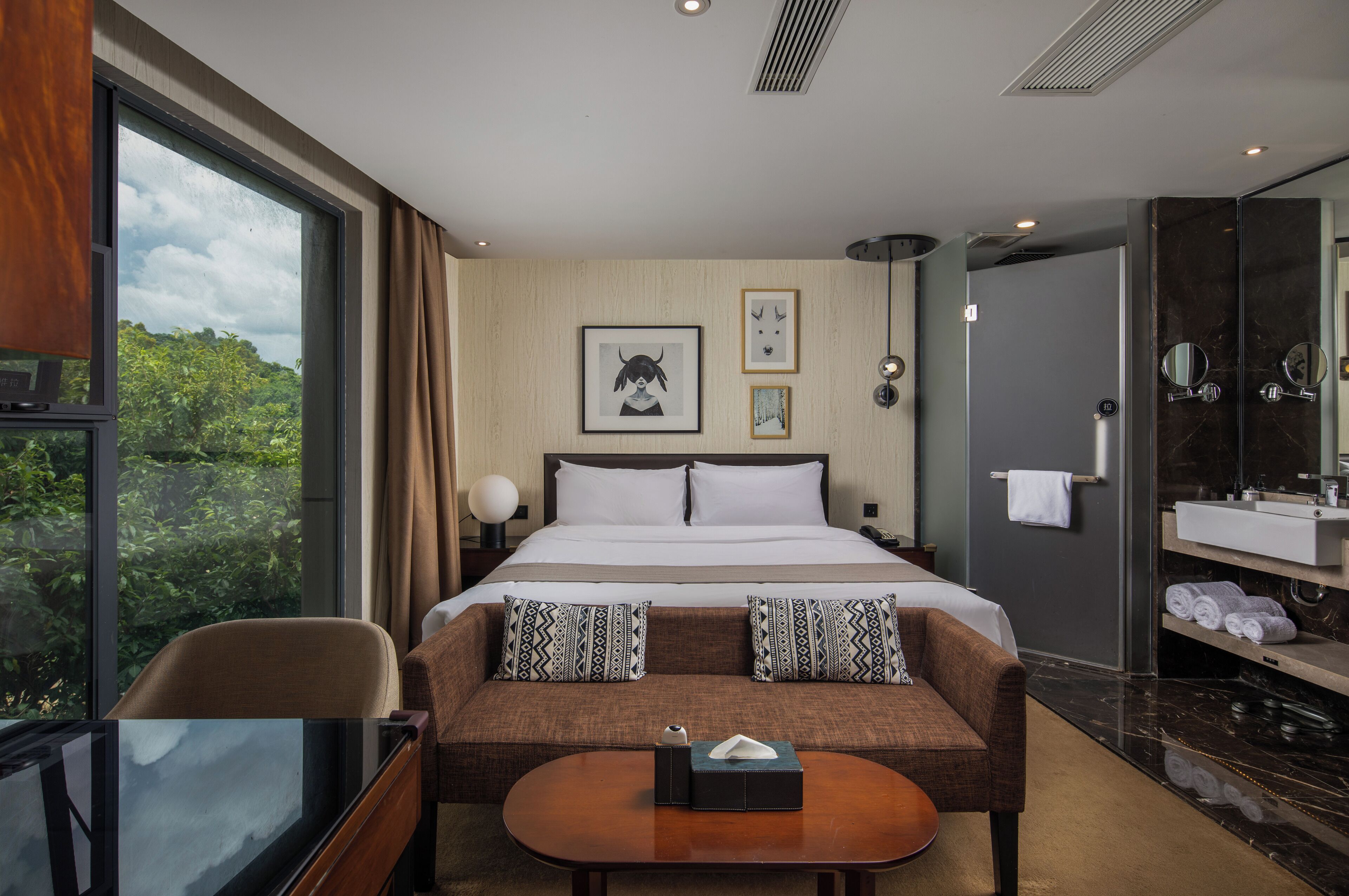 A hotel room with a large bed, a sofa, a small table, a bathroom with a mirror and sink, and a view of greenery through a window.