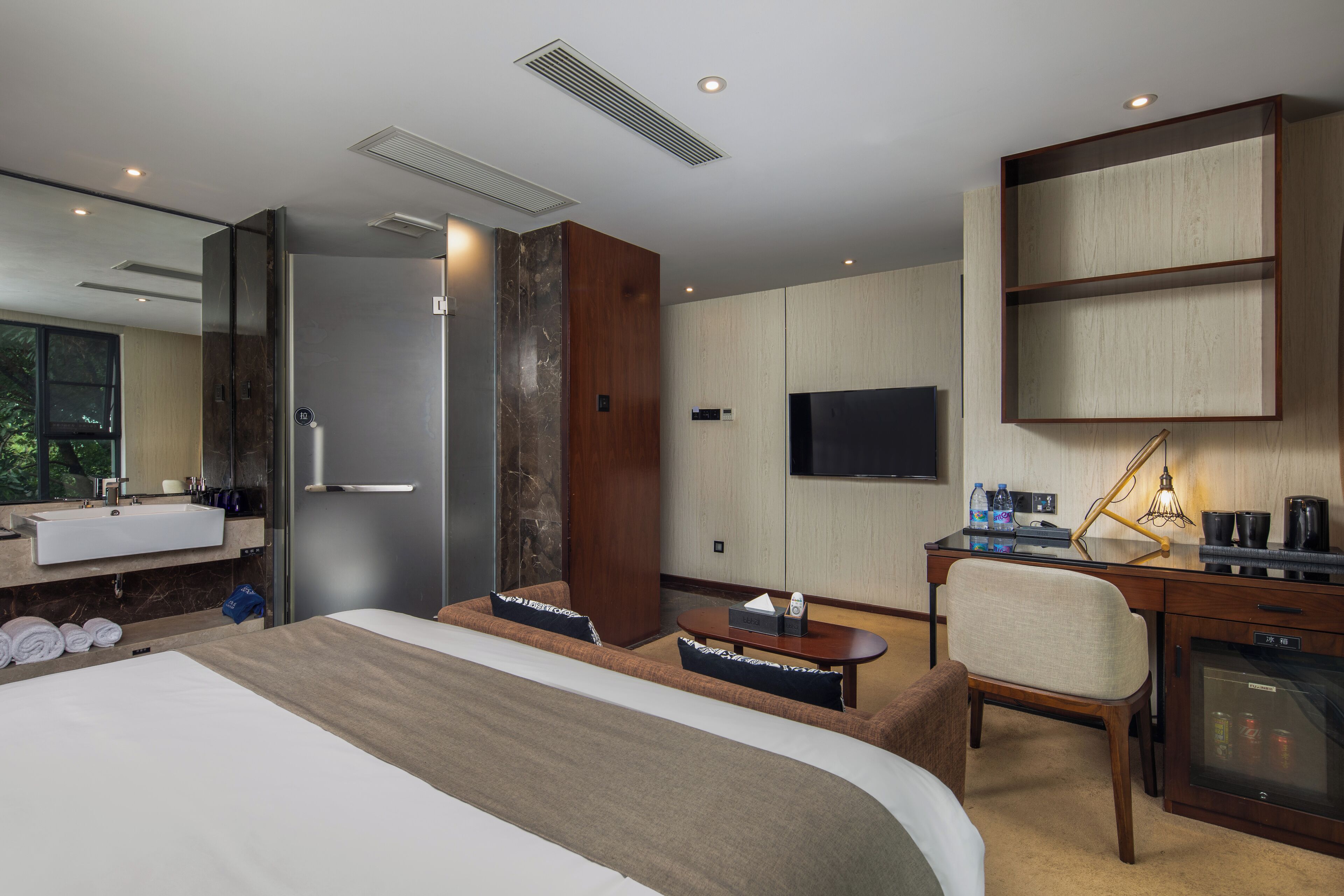 A modern hotel room with a large bed, a desk, a chair, a TV, a minibar, and a bathroom with a sink and shower.