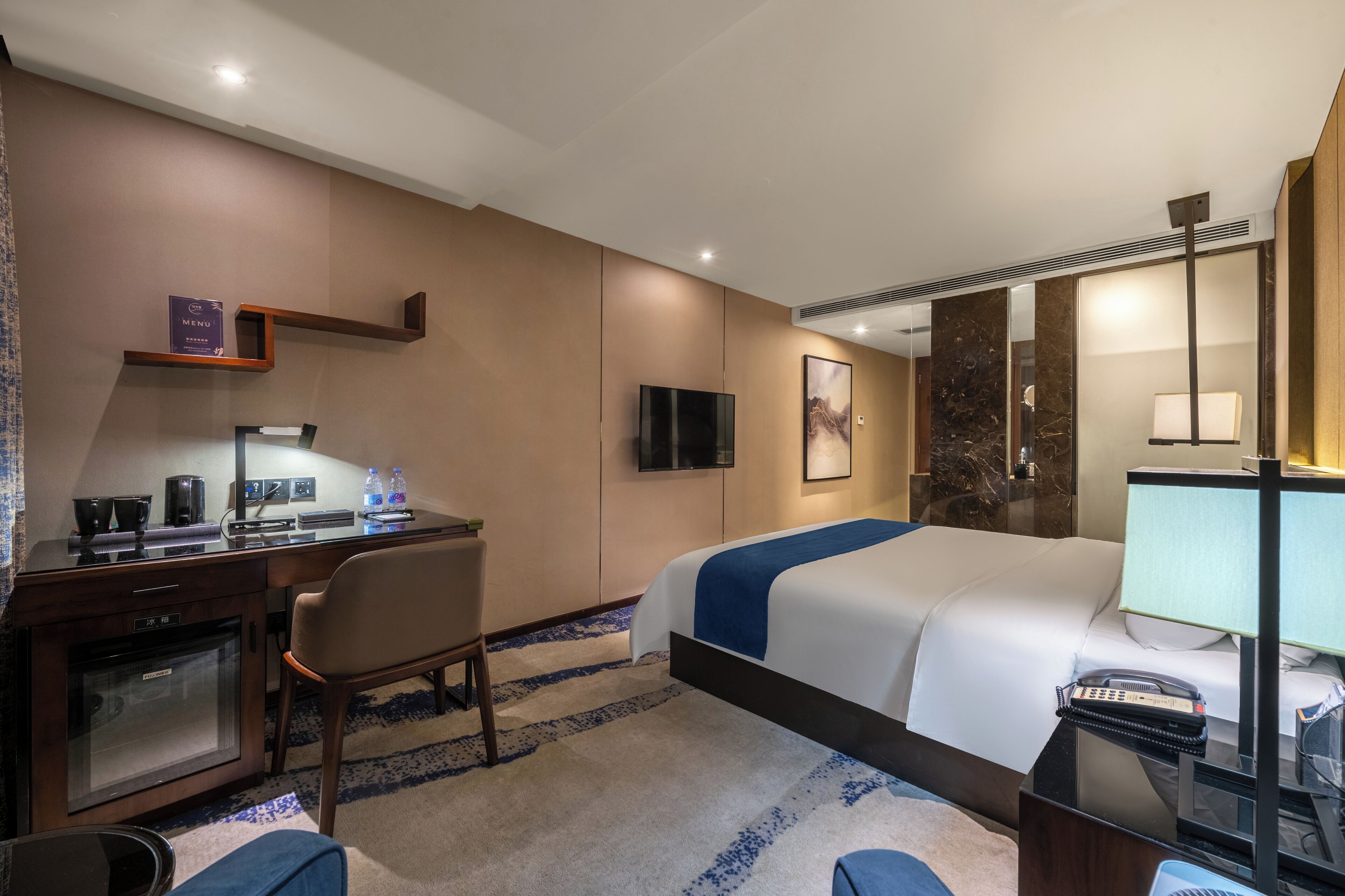 A hotel room with a bed, desk, chair, TV, and a minibar.