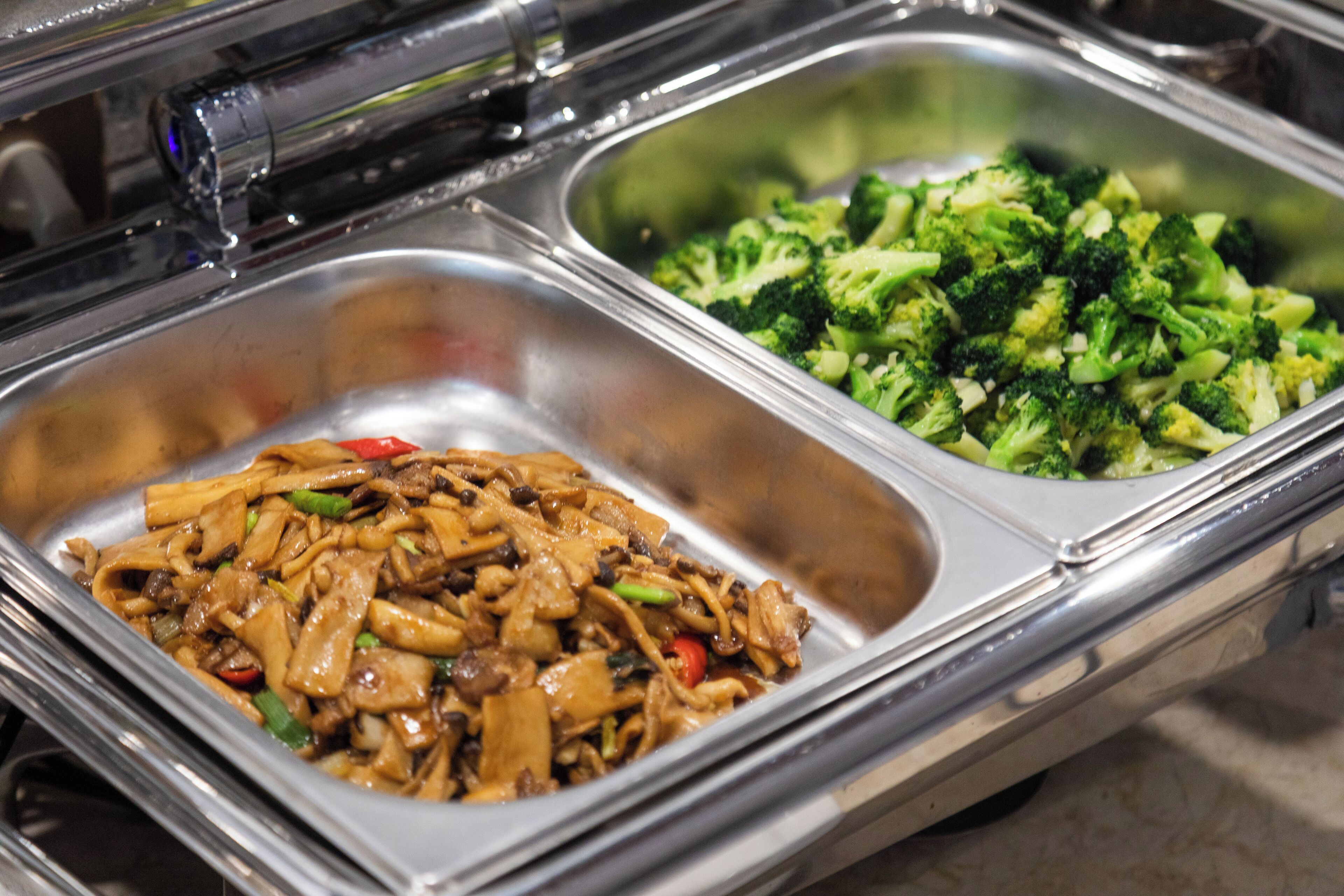 Buffet com duas bandejas de metal, uma com noodles refogados e outra com brócolis cozido no vapor.