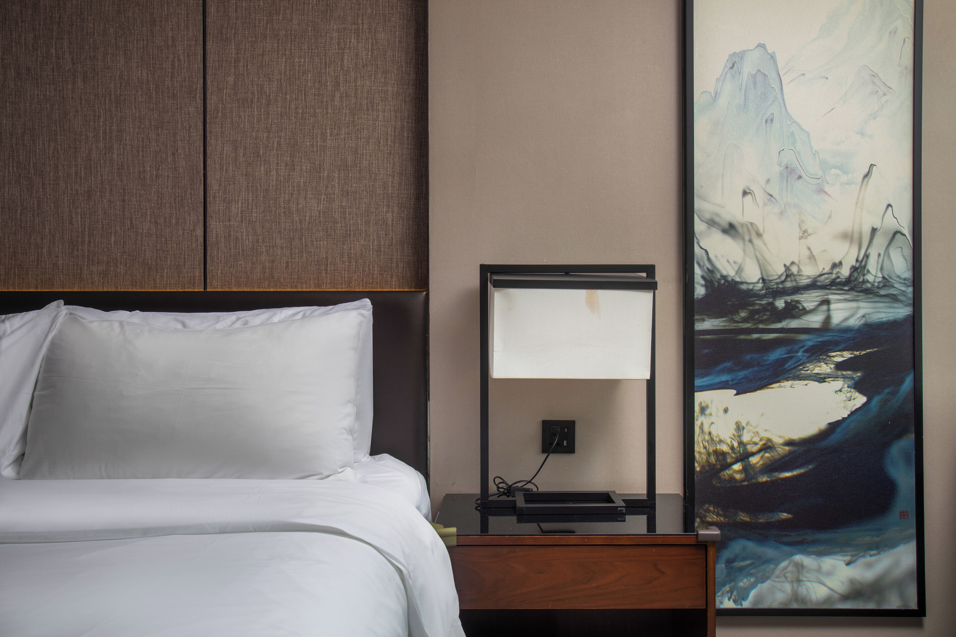 A neatly made bed with white linens, a bedside table with a lamp, and a large abstract painting on the wall.