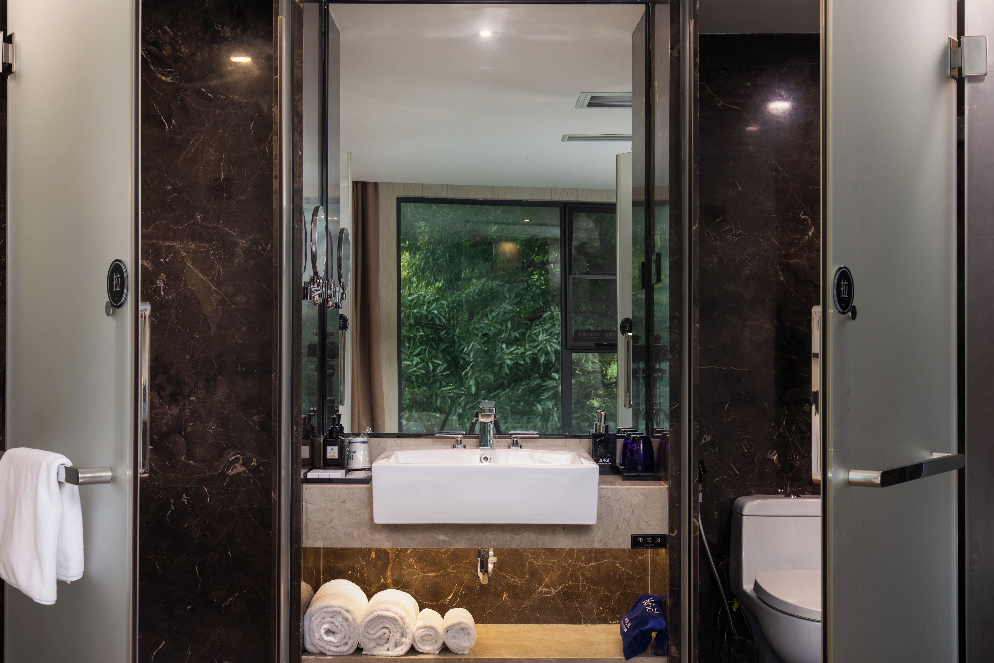 A modern bathroom with a large mirror, a white sink, and a toilet. There are towels on a shelf and a view of greenery outside.