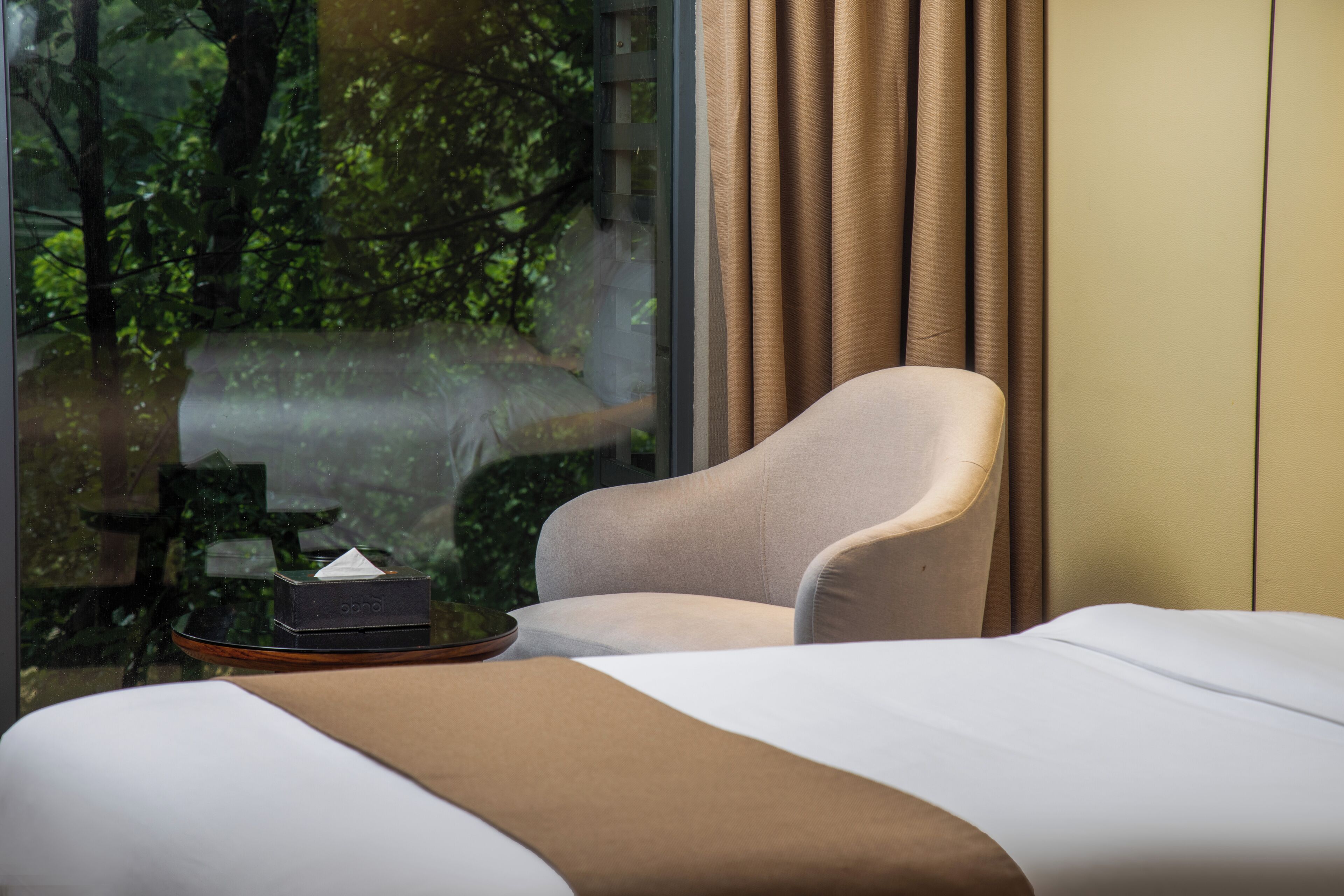 A hotel room with a bed, a chair, a table with a tissue box, and a view of greenery through a window.