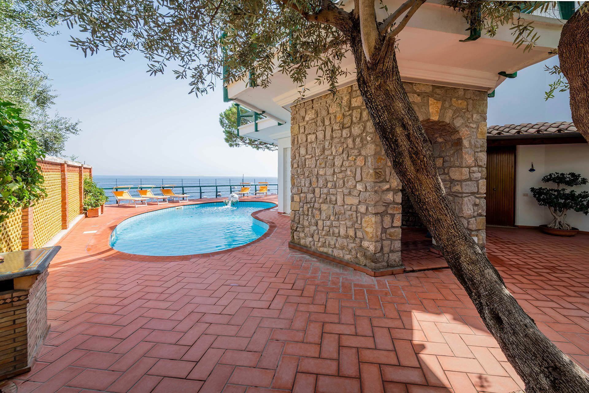 A pool area with a stone wall, a tree, and a view of the sea.