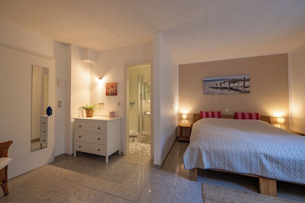 A bedroom with a bed, bedside tables, a nightstand, a dresser, a mirror, and a bathroom visible through an open door.