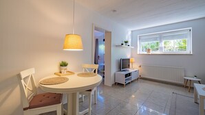 Ein modernes Wohnzimmer mit einem runden Esstisch, weißen Stühlen, einem Fernseher und einem Fenster mit Blick auf Grünflächen.
