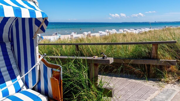 Blick auf einen Strand mit einer Holzterrasse, gestreiften Strandsonnenschirmen und Liegestühlen.