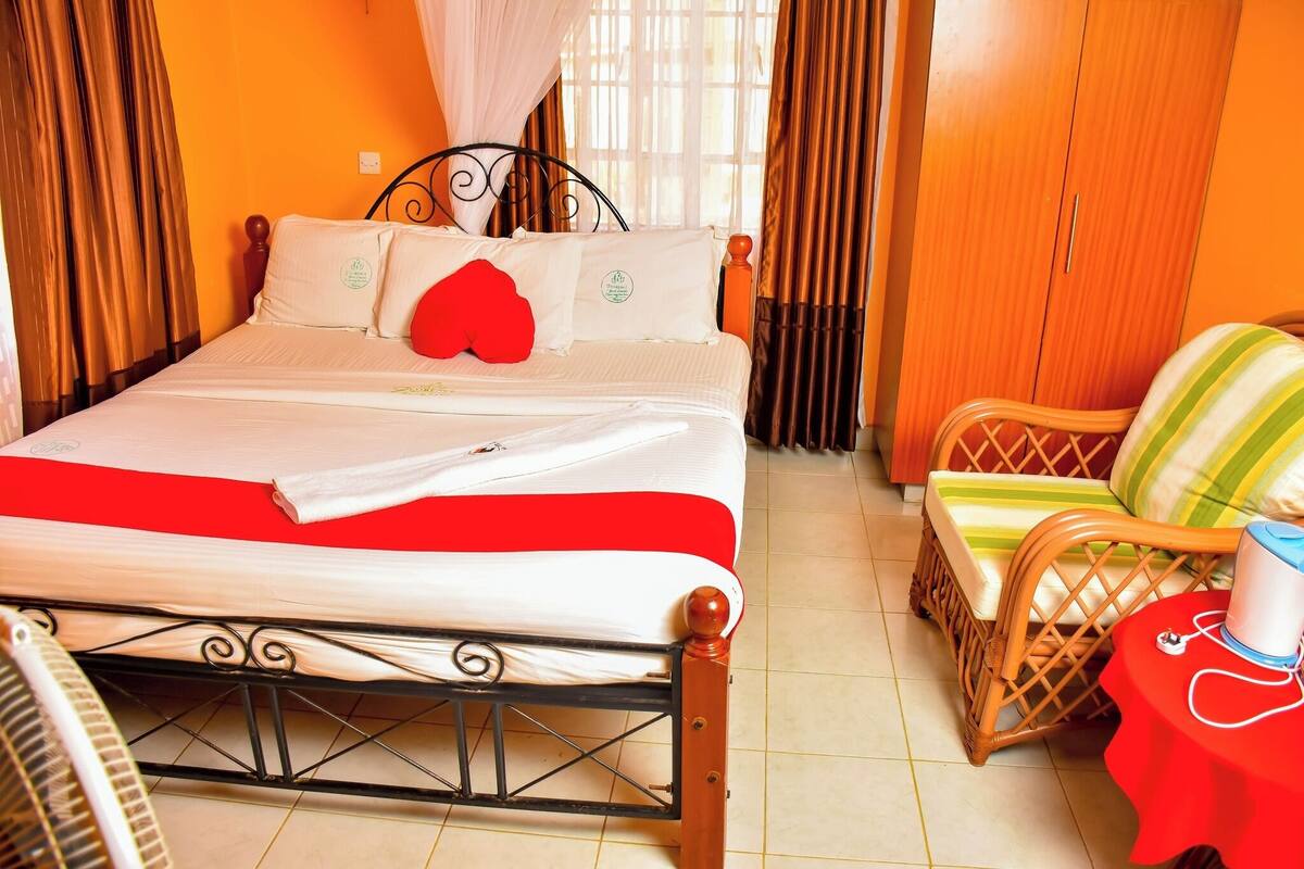 A neatly made bed with a red and white bedspread, a wooden headboard, and a red pillow. There is a wicker chair with a striped cushion and a small table with a red tablecloth and a cup.