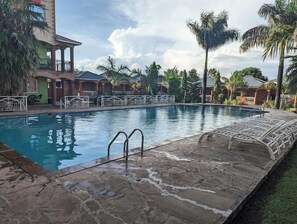 A swimming pool with stone flooring, surrounded by palm trees and a building with a green facade. - Florence Hotel (Migori)