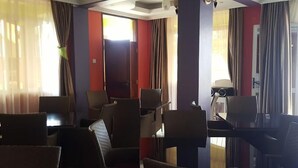 Ένα δωμάτιο ξενοδοχείου με καθιστικό που περιλαμβάνει πολλές καρέκλες και ένα τραπέζι, μεγάλα παράθυρα με κ ουρτίνες και μια πόρτα στο βάθος.