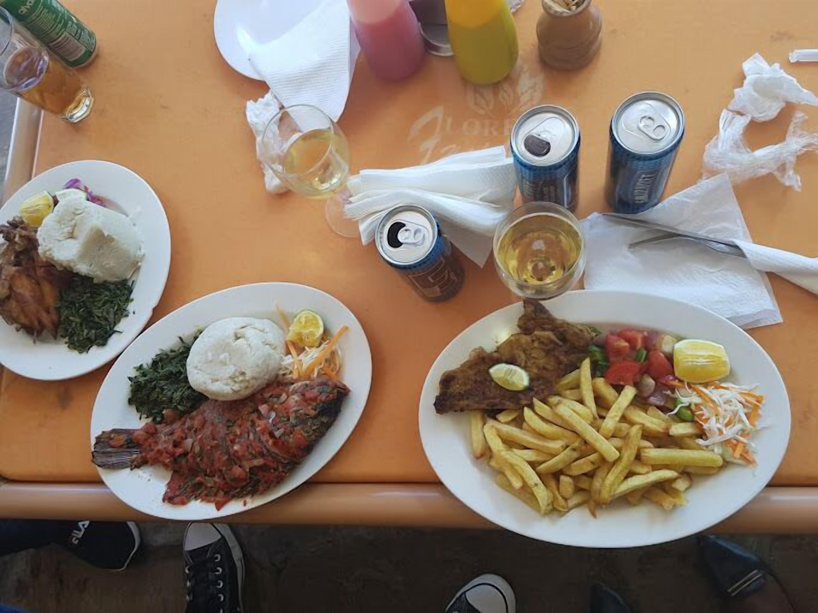 A table with two plates of food, a glass of wine, and several cans of soda.