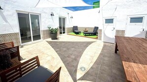A covered outdoor patio with a wooden table and chairs, a small seating area with lounge chairs, and a potted plant.