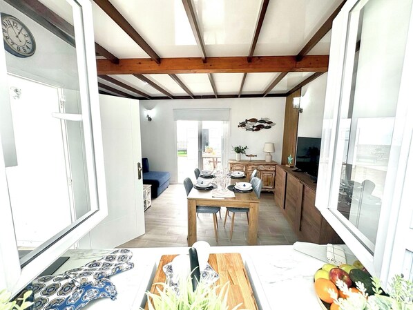 Una habitación luminosa con techo de madera, una mesa de comedor lista para cuatro personas y una cocina visible a través de una puerta abierta.