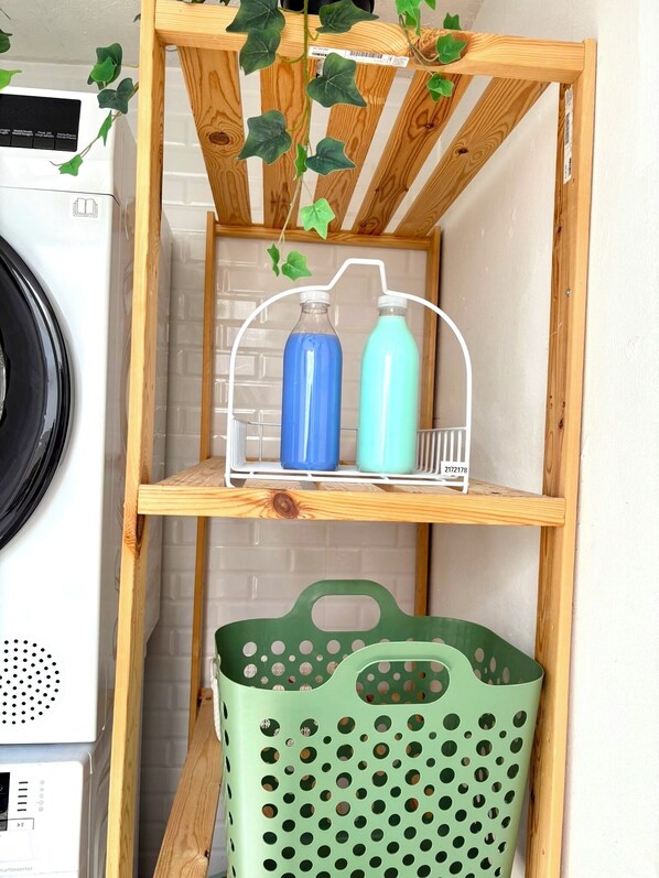Una estantería de madera con una planta colgante, un escurridor metálico que sostiene dos botellas y una cesta verde en la parte inferior.