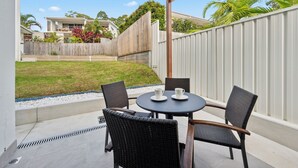 Une terrasse couverte avec une table ronde et quatre chaises, donnant sur un jardin avec une clôture en bois et une maison en arrière-plan.