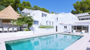 Una villa blanca moderna con piscina, mobiliario exterior y una estructura de techo de paja.